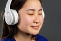 Young beautiful asian woman with headphones in modern wear enjoying and dancing on grey background. Radio, melody