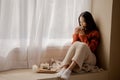 Young beautiful Asian woman holding cup of coffee and marshmallows on top, sitting at home and looking out the window Royalty Free Stock Photo