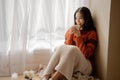 Young beautiful Asian woman holding cup of coffee and marshmallows on top, sitting at home and looking out the window Royalty Free Stock Photo