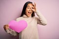 Young beautiful asian woman holding pink heart standing over isolated background with happy face smiling doing ok sign with hand Royalty Free Stock Photo