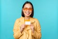 Young beautiful asian woman showing credit card, smiling, choosing bank, standing over blue background