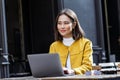 Young beautiful Asian woman sitting in coffee shop at wooden table, drinking coffee and using laptop. Girl browsing internet, Royalty Free Stock Photo