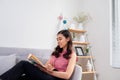 Young beautiful asian woman sitting on couch reading a book enjoying her tea in living room at home Royalty Free Stock Photo