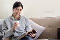 Young beautiful asian woman student sitting on sofa reading book at home while using laptop for online learning Royalty Free Stock Photo