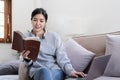 Young beautiful asian woman student sitting on sofa reading book at home while using laptop for online learning Royalty Free Stock Photo