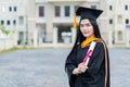 A young beautiful Asian woman university graduate in graduation gown and mortarboard holds a degree certificate stands in front of Royalty Free Stock Photo