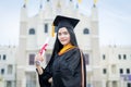 A young beautiful Asian woman university graduate in graduation gown and mortarboard holds a degree certificate stands in front of Royalty Free Stock Photo