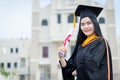 A young beautiful Asian woman university graduate in graduation gown and mortarboard holds a degree certificate stands in front of Royalty Free Stock Photo
