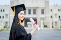 A young beautiful Asian woman university graduate in graduation gown and mortarboard holds a degree certificate stands in front of Royalty Free Stock Photo