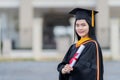 A young beautiful Asian woman university graduate in graduation gown and mortarboard holds a degree certificate stands in front of Royalty Free Stock Photo