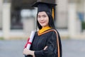 A young beautiful Asian woman university graduate in graduation gown and mortarboard holds a degree certificate stands in front of Royalty Free Stock Photo