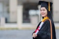 A young beautiful Asian woman university graduate in graduation gown and mortarboard holds a degree certificate stands in front of Royalty Free Stock Photo
