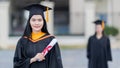 A young beautiful Asian woman university graduate in graduation gown and mortarboard holds a degree certificate stands in front of Royalty Free Stock Photo