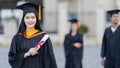 A young beautiful Asian woman university graduate in graduation gown and mortarboard holds a degree certificate stands in front of Royalty Free Stock Photo