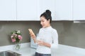 Young beautiful Asian woman using phone and drinking milk while standing in kitchen Royalty Free Stock Photo
