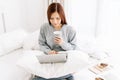 Young beautiful Asian woman in warm knitted clothes with cup of coffee and working on a laptop sitting on the bed in the house. Royalty Free Stock Photo