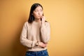 Young beautiful asian woman wearing casual sweater over yellow isolated background with hand on chin thinking about question,