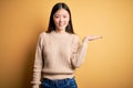 Young beautiful asian woman wearing casual sweater over yellow isolated background smiling cheerful presenting and pointing with Royalty Free Stock Photo