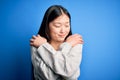 Young beautiful asian woman wearing casual sweater standing over blue isolated background Hugging oneself happy and positive, Royalty Free Stock Photo