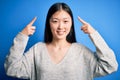 Young beautiful asian woman wearing casual sweater standing over blue isolated background smiling pointing to head with both hands Royalty Free Stock Photo