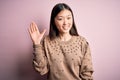 Young beautiful asian woman wearing fashion and elegant sweater over pink solated background Waiving saying hello happy and
