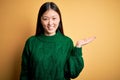 Young beautiful asian woman wearing green winter sweater over yellow isolated background smiling cheerful presenting and pointing Royalty Free Stock Photo