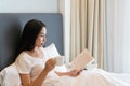 Young beautiful Asian woman in white T-shirt pajamas drinking coffee while reading a book sitting on the bed in the morning. Copy Royalty Free Stock Photo