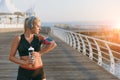 Young beautiful athletic girl with long blond hair in headphones and a bottle of water in hands looks at mobile phone on hand by t Royalty Free Stock Photo