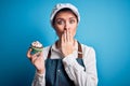 Young beautiful baker woman with blue eyes wearing apron holding chocolate cupcake cover mouth with hand shocked with shame for Royalty Free Stock Photo