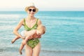 Young beautiful bald mother carrying cute caucasian toddler boy out clear blue water at tropical beach on bright sunny day. Mom