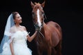 Young beautiful beauty bride in fashion white wedding costume stand with handsome horse on black background Royalty Free Stock Photo