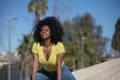 Young, beautiful, black woman with afro hair, wearing yellow t-shirt and jeans, sitting astride a stone wall, smiling, happy and Royalty Free Stock Photo
