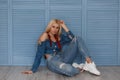 Young beautiful blond girl model in fashion denim clothes with stylish jeans and white sneakers sitting near the blue wooden wall.