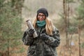 Young beautiful blond woman in camouflage outfit and green scarf posing with thermos in the forest. Travel lifestyle concept.