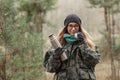 Young beautiful blond woman in camouflage outfit and green scarf posing with thermos in the forest. Travel lifestyle concept.