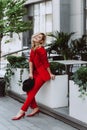 Young beautiful blond woman in a red suit and black hat stands outdoors in the yard next to the building. Stylish people. Soft Royalty Free Stock Photo