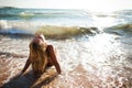 Young beautiful blond woman in white bikini sitting on sea water edge in waves foam and enjoying sunshine