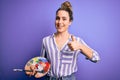 Young beautiful blonde artist woman painting using brush and palette over purple background happy with big smile doing ok sign, Royalty Free Stock Photo