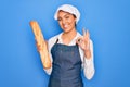 Young beautiful blonde baker woman with blue eyes holding loaf of homemade fresh bread doing ok sign with fingers, excellent Royalty Free Stock Photo