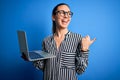 Young beautiful blonde businesswoman wearing glasses using laptop over blue background pointing and showing with thumb up to the Royalty Free Stock Photo