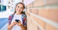 Young beautiful blonde caucasian woman smiling happy outdoors on a sunny day wearing headphones and using smartphone leaning on a Royalty Free Stock Photo