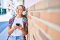 Young beautiful blonde caucasian woman smiling happy outdoors on a sunny day wearing headphones and using smartphone leaning on a Royalty Free Stock Photo