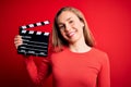 Young beautiful blonde clapper woman holding clapboard over isolated red background with a happy face standing and smiling with a Royalty Free Stock Photo