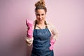 Young beautiful blonde cleaner woman doing housework wearing arpon and gloves angry and mad raising fist frustrated and furious Royalty Free Stock Photo