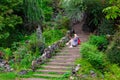 Young beautiful blonde girl in a blue raincoat sits on a wooden staircase with stone walls in a green flowered garden texting on Royalty Free Stock Photo