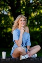 Young beautiful blonde girl in a blue short dress and with a white belt bag walks in a green park. He sits on a bench Royalty Free Stock Photo