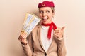 Young beautiful blonde girl wearing stewardess uniform holding boarding pass pointing thumb up to the side smiling happy with open