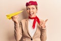 Young beautiful blonde girl wearing stewardess uniform holding paper plane pointing thumb up to the side smiling happy with open