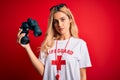 Young beautiful blonde lifeguard woman wearing t-shirt with red cross using binoculars with a confident expression on smart face