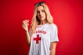 Young beautiful blonde lifeguard woman wearing t-shirt with red cross using whistle with a confident expression on smart face